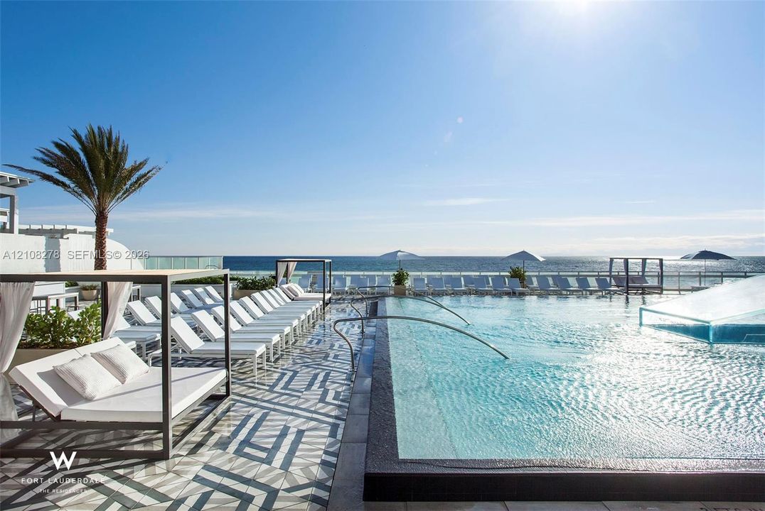 Infinity-edge swimming pool featuring a patterned tile deck, metal-framed structures, and a glass-paneled perimeter railing