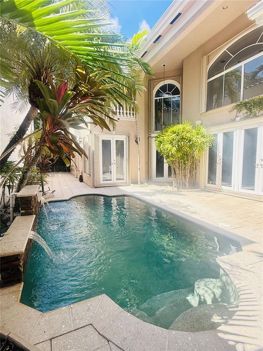 Exterior courtyard featuring a custom swimming pool with integrated water features, stone paver decking, multiple arched glass entryways, and two-story stucco architecture