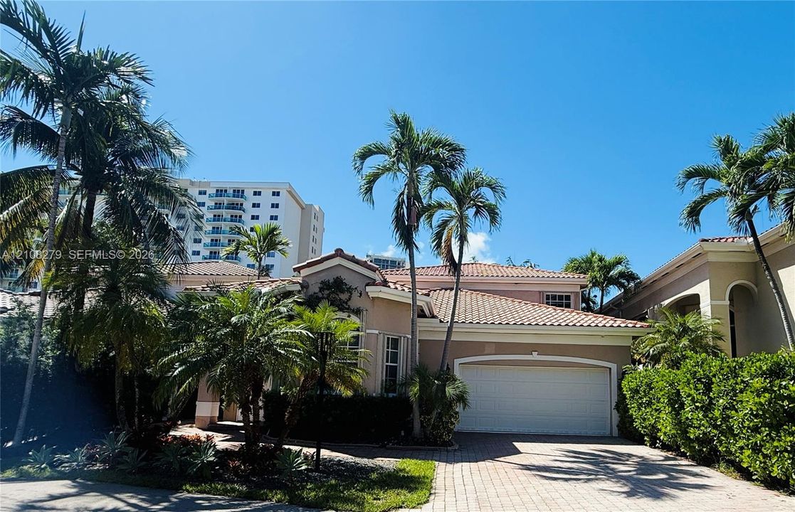 Stucco exterior residence featuring a multi-pitched clay tile roof, arched garage entrance, and paved driveway surrounded by mature palm trees and manicured hedging