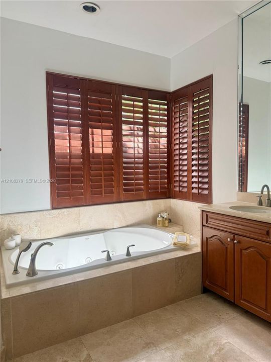 Jetted soaking tub with deck-mounted chrome fixtures, custom wood shutters, stone-tiled tub surround, wood vanity cabinetry, and large-format tile flooring