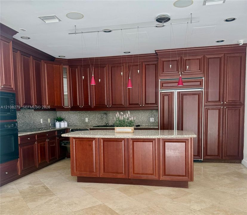 Custom wood-finish cabinetry, central island with stone countertop, integrated refrigerator, stone tile flooring, and recessed ceiling lighting