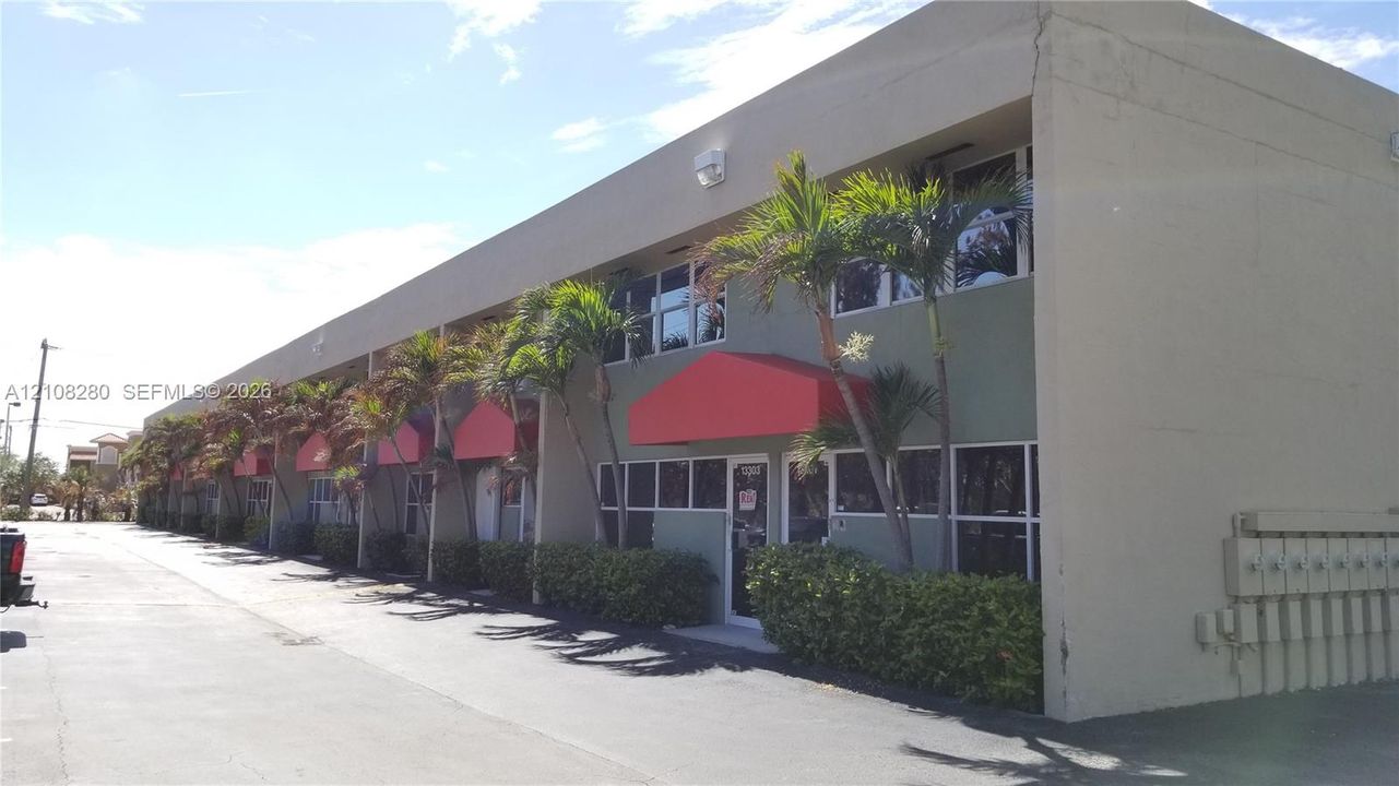 Multi-unit building exterior featuring concrete construction, red fabric awnings, large-pane glass windows, and paved asphalt parking