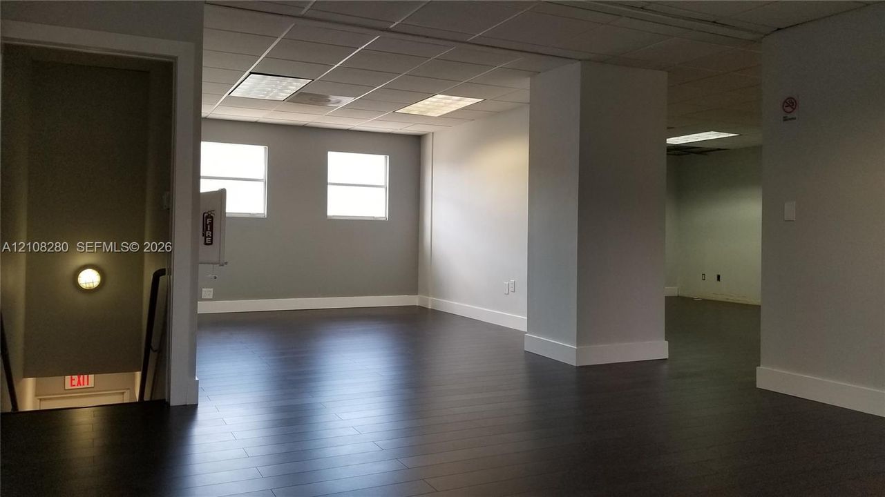 Open-concept interior featuring dark wood-finish flooring, neutral wall tones, recessed drop-ceiling grid lighting, and rectangular windows