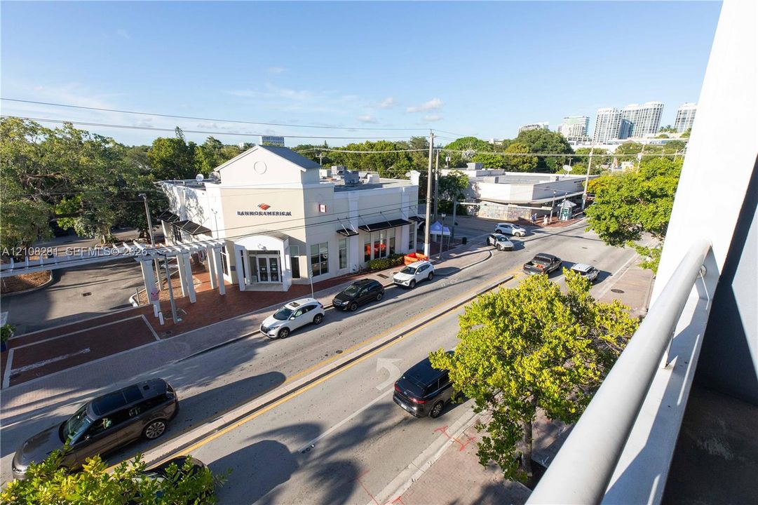 Elevated balcony perspective overlooking a streetscape featuring mature trees, paved roadways, and surrounding commercial structures