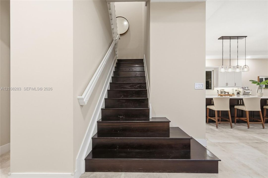 Staircase featuring dark wood-finish treads, white handrail, and white balusters, situated on stone-finish flooring with a multi-pendant light fixture and built-in cabinetry in the background