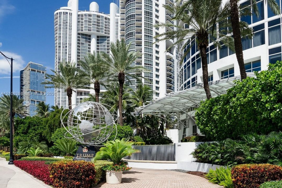 High-rise building exterior featuring a white curved facade, metal-frame porte-cochère, decorative metal globe sculpture, tiered water feature, and mature tropical landscaping
