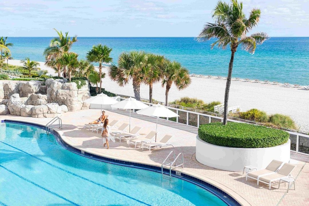 Curved swimming pool featuring a stone-faced grotto, expansive paver pool deck, elevated circular planter with mature palm tree, and white perimeter fencing
