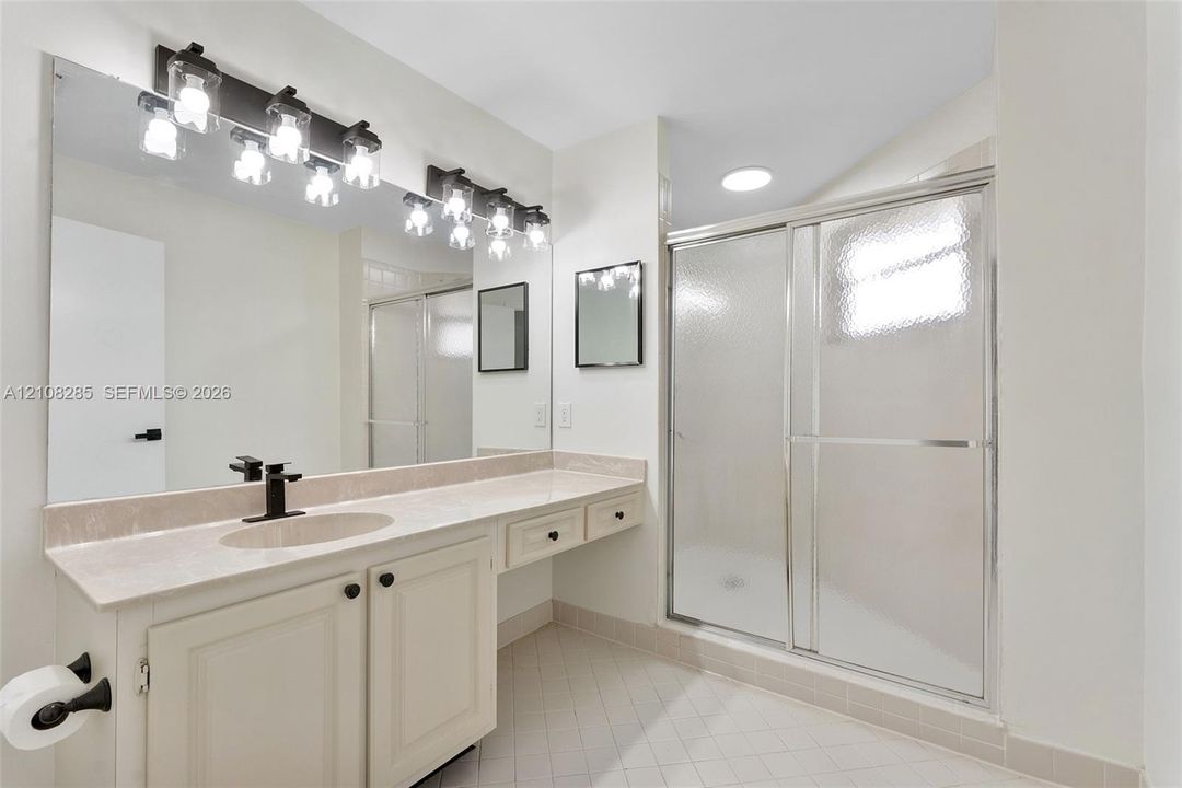 Bathroom featuring an expansive vanity with stone-style countertop, undermount sink with matte black faucet, wall-to-wall mirror, multi-light fixture, and glass-enclosed shower