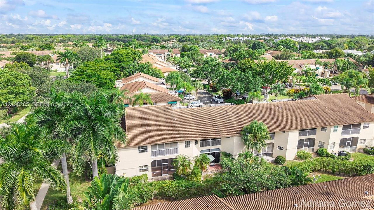 Multi-story residential building with a tile roof, light-colored stucco exterior, and screened-in lanais, set within a landscape of mature palm trees and greenery