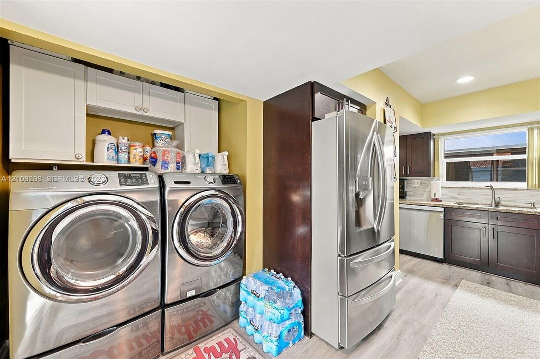 Laundry area featuring stainless steel front-load appliances and custom white cabinetry, incorporating a kitchen space with dark-toned cabinetry, stainless steel refrigerator and dishwasher, and stone-surfaced countertops