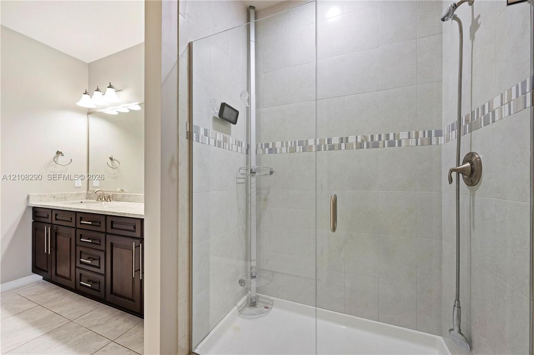 Walk-in shower with a glass enclosure, tile surround, and metallic fixtures, featuring a vanity with stone-finish countertop, dark wood cabinetry, and a full-width wall mirror