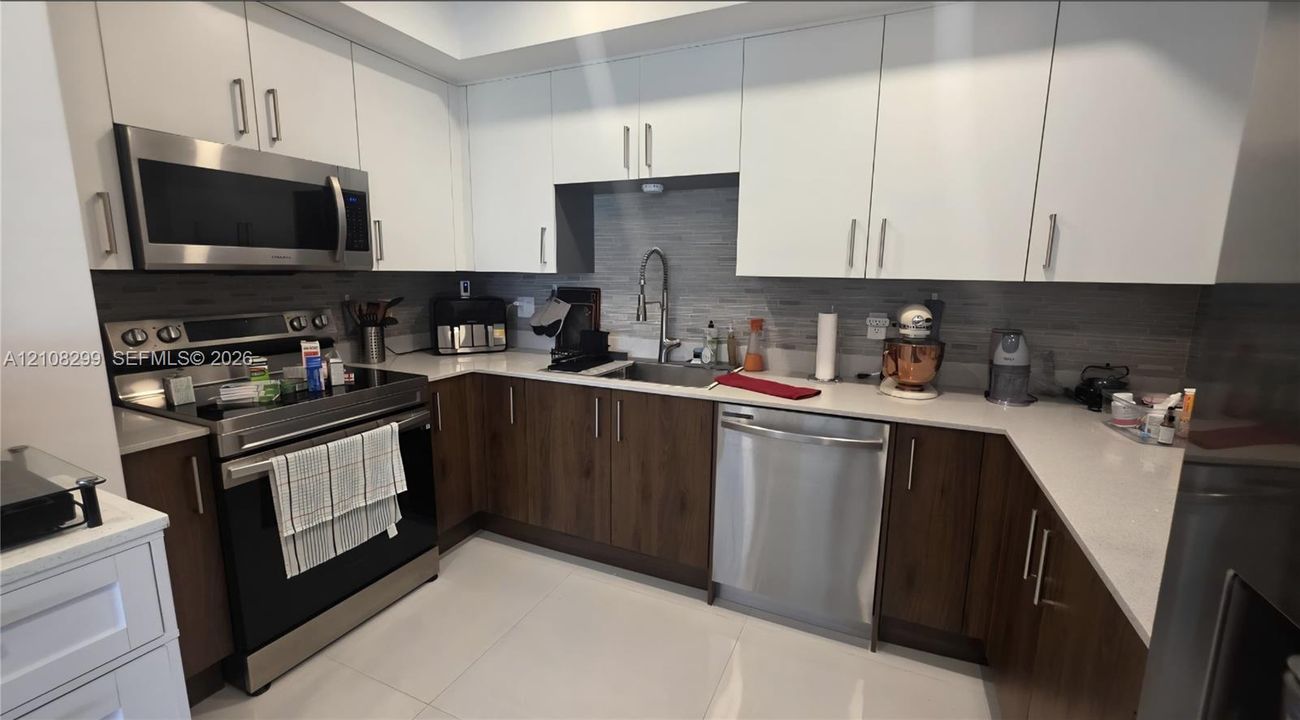 Kitchen featuring two-tone cabinetry with upper white cabinets and lower wood-finish cabinets, light-toned stone-composite countertops, stainless steel appliances, and a linear tile backsplash