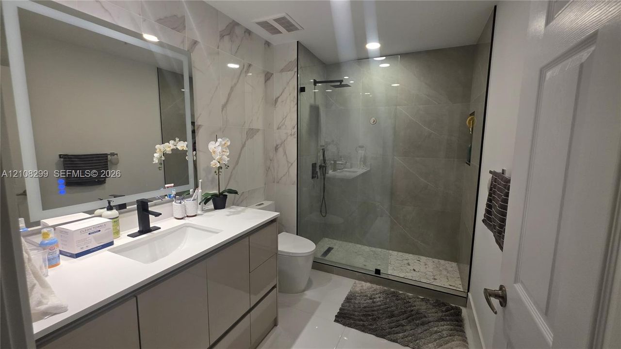 Modern bathroom featuring a high-gloss vanity with a white countertop, wall-mounted illuminated mirror, and a walk-in shower with a glass enclosure, rainfall showerhead, and stone-finish floor tiling
