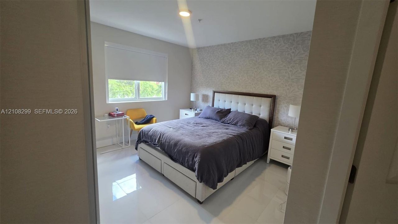 Bedroom featuring light-toned tile flooring, neutral wall paint, patterned accent wall covering, and a window with a retractable roller shade