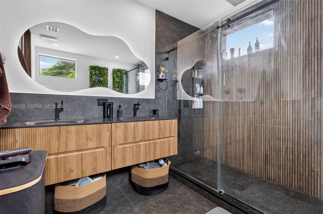 Floating wood-finish vanity with dual sinks, backlit organic-shaped mirror, matte black fixtures, walk-in shower with glass enclosure, and vertical-slatted tile shower wall