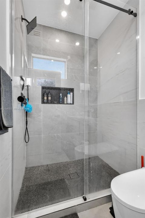 Walk-in shower featuring large-format light grey wall tile, a textured pebble-style floor, a built-in recessed niche, a frameless glass enclosure, and a matte black rainfall showerhead