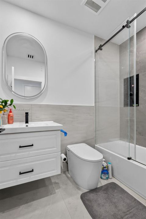 Bathroom featuring a white wall-mounted vanity with black hardware, an oval mirror, a tub-shower combination with a glass sliding enclosure, and large-format neutral-tone tile