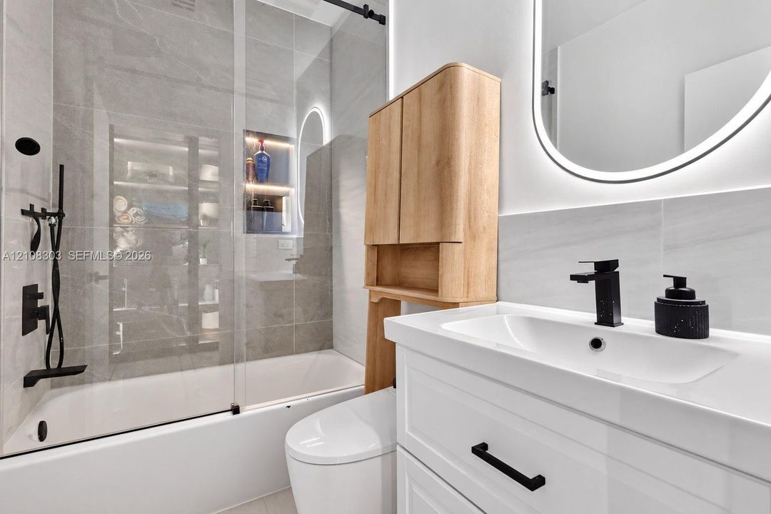Bathroom featuring a glass-enclosed shower with large-format tile surround, recessed niche lighting, white vanity cabinet with matte black hardware, integrated vanity mirror with LED illumination, and wood-finish cabinetry