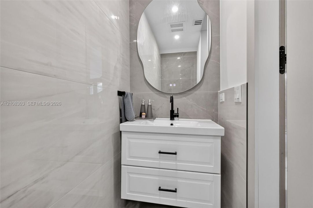 Wall-mounted white vanity with two drawers, rectangular basin, matte black faucet, and large organic-shaped mirror, featuring floor-to-ceiling large-format tile surround