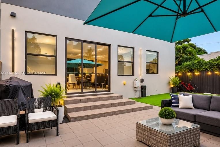 Patio area featuring light-toned tile flooring, three-step tiered entry, sliding glass doors, vertical exterior wall lighting, and an artificial grass lawn