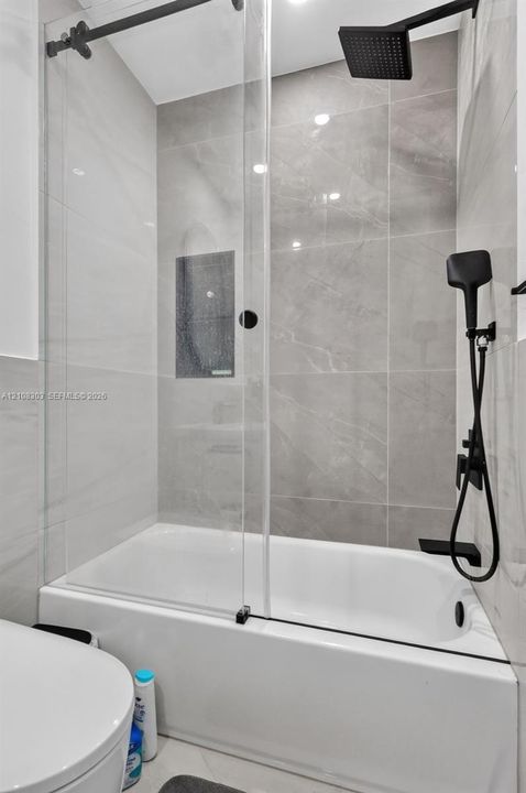Shower and bathtub combination featuring large-format stone-finish tile surround, glass panel enclosure, matte black rainfall showerhead, and matching matte black handheld fixture