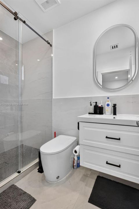 Walk-in shower with a sliding glass door, white wall-mounted vanity with two drawers, matte black hardware, oval-framed mirror, and large-format tile flooring