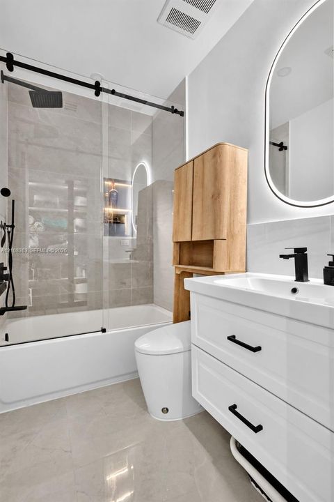 Bathroom featuring a white two-drawer vanity with matte black hardware, a shower-tub combination with a glass sliding door, large-format tile surround, an illuminated oval mirror, and a built-in wood-finish cabinet