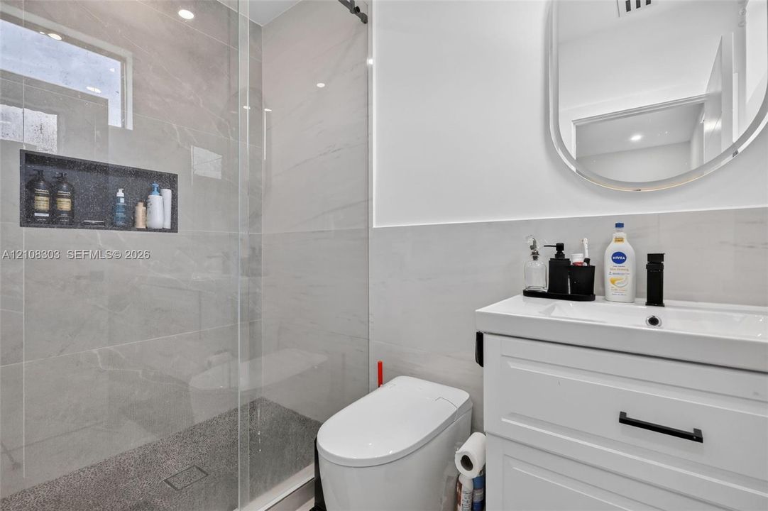 Walk-in shower with frameless glass enclosure, built-in tiled niche, white vanity with rectangular sink, matte black faucet, and oval wall-mounted mirror