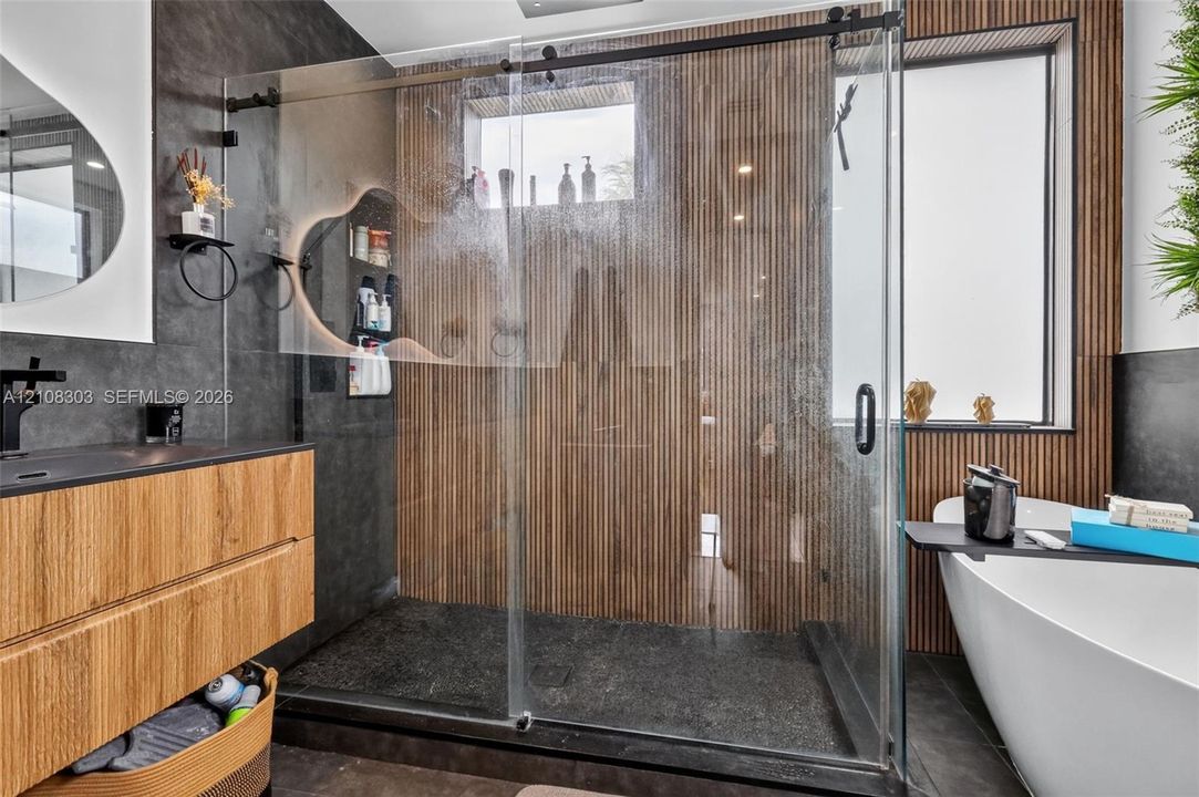 Walk-in shower featuring a frameless glass enclosure, floor-to-ceiling slatted wood-finish wall panels, large-format dark tile surround, and a freestanding soaking tub