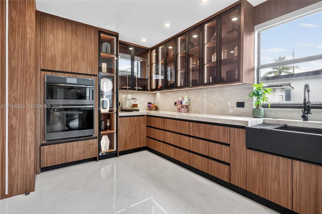 Wood-finish cabinetry with glass-front display panels, integrated double wall ovens, stone-finish countertops, matte black apron-front sink, and polished tile flooring