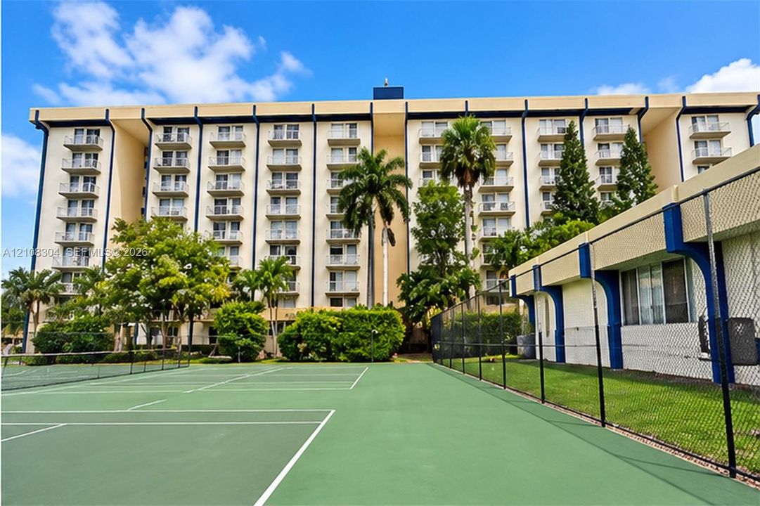 Multi-story residential building featuring private balconies, tennis courts, chain-link perimeter fencing, and established palm trees