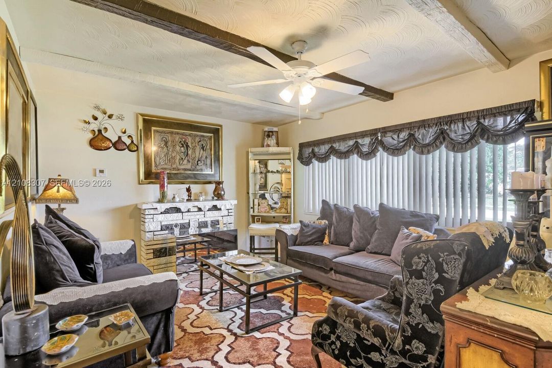 Living area featuring exposed wooden ceiling beams, patterned ceiling detail, stone-faced fireplace, and a large window with horizontal blinds