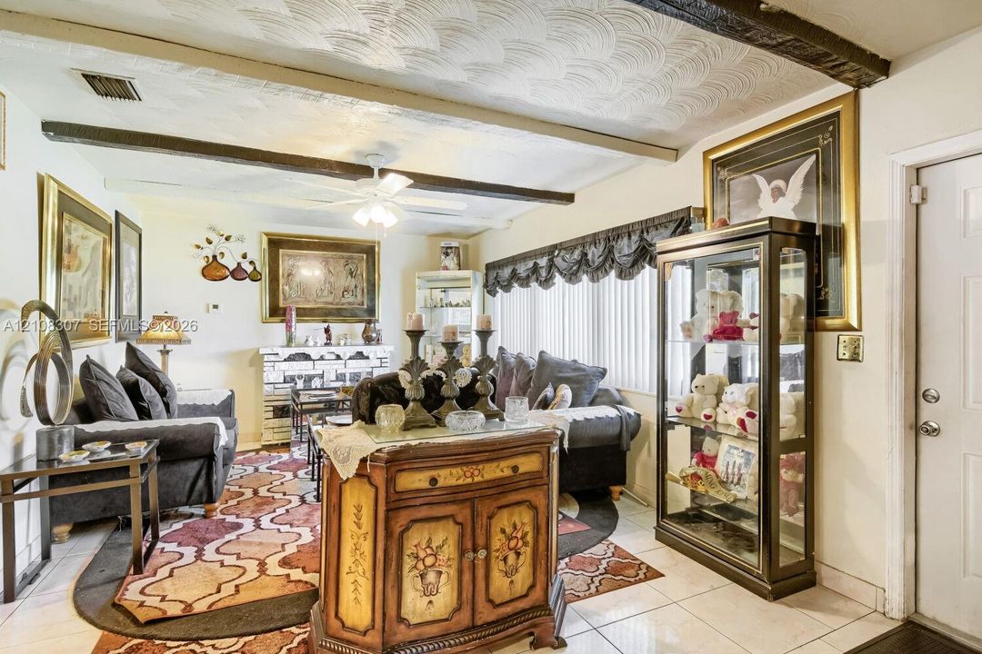 Living area featuring exposed ceiling beams, textured ceiling finish, ceramic tile flooring, built-in masonry fireplace, and ceiling fan