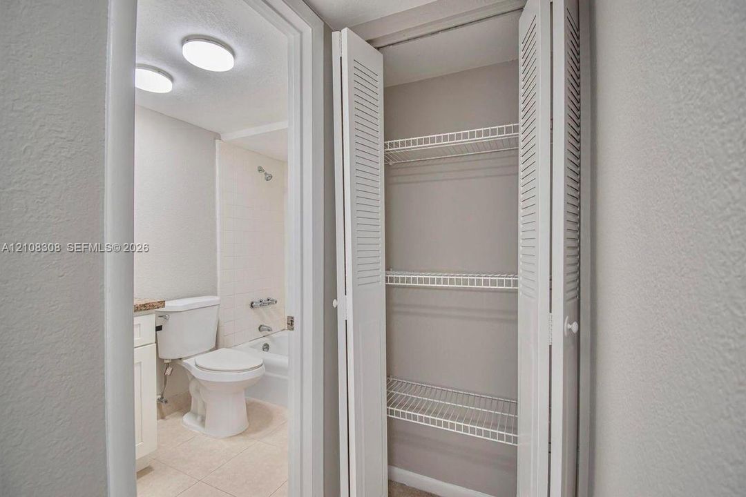Built-in storage closet with louvered bi-fold doors and wire shelving, featuring a tiled bathtub enclosure, porcelain toilet, and recessed ceiling lighting