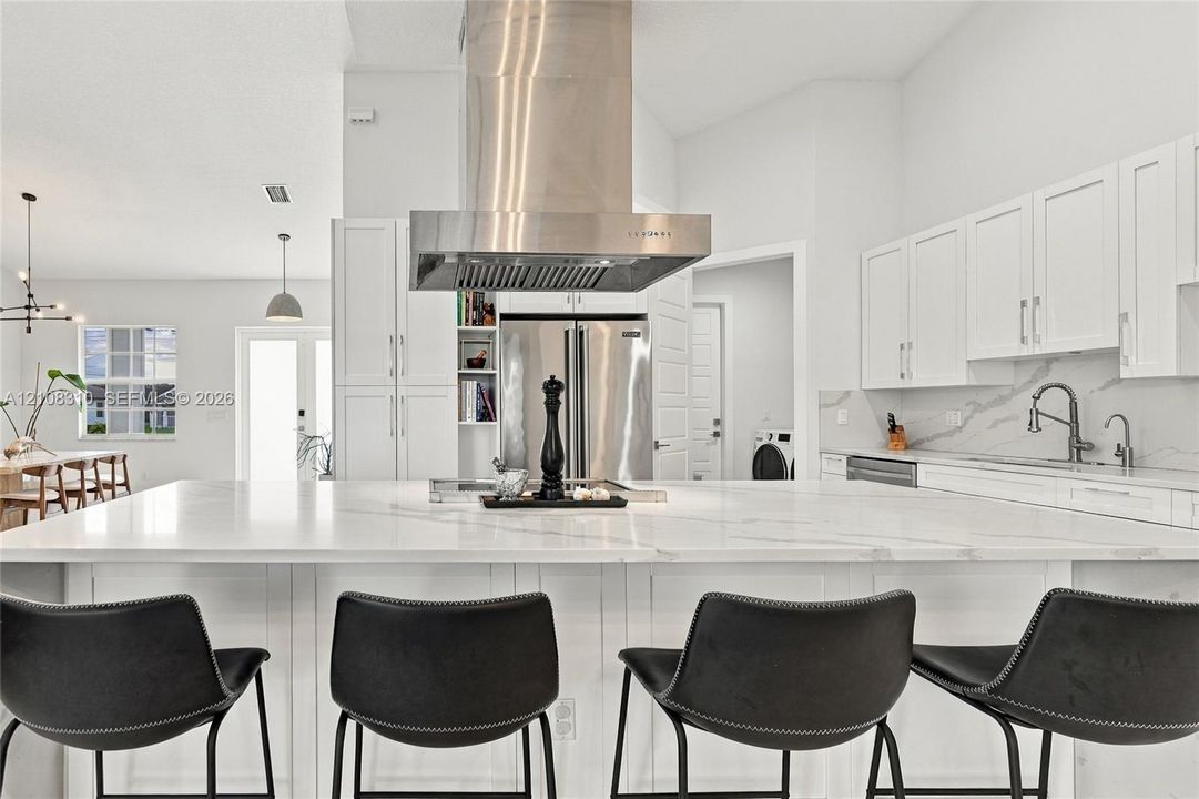 Kitchen featuring a large central island with solid-surface countertop, white shaker-style cabinetry, stainless steel range hood, and integrated stainless steel appliances