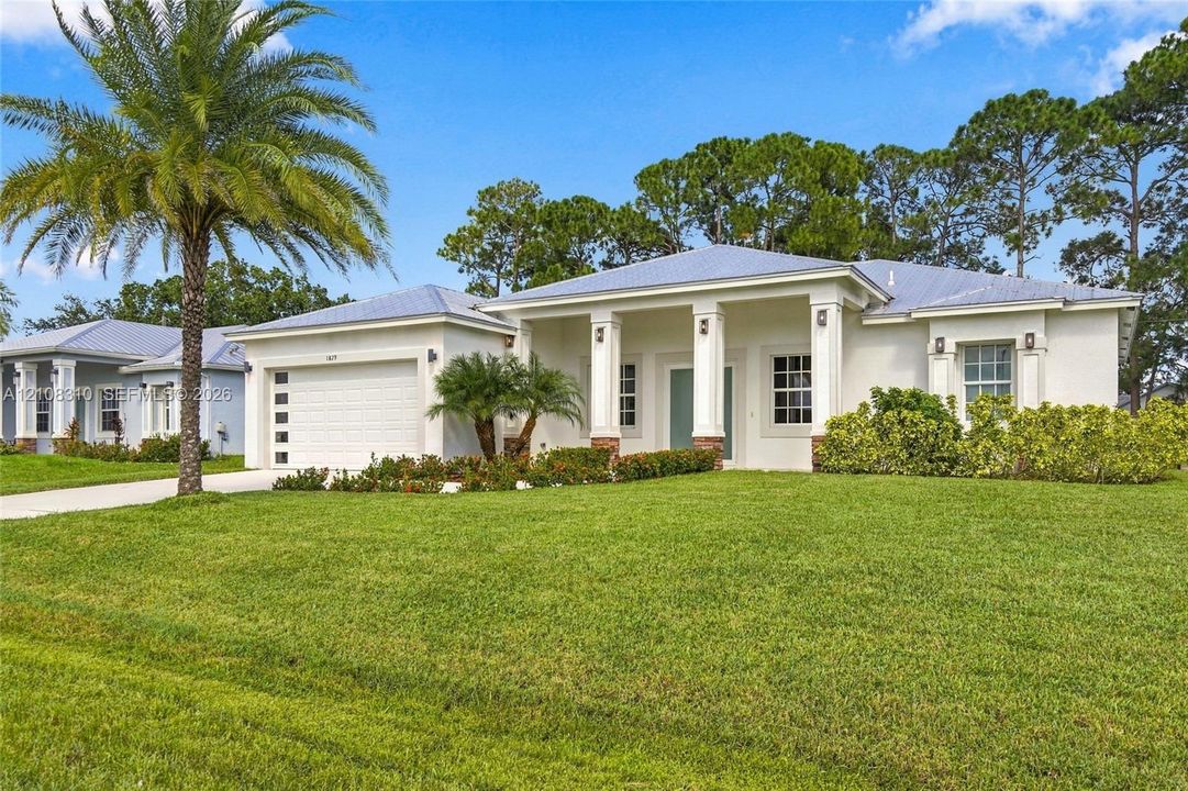 Single-story exterior featuring a metal roof, white stucco facade, covered front entryway with square columns, attached multi-car garage, and manicured landscaping