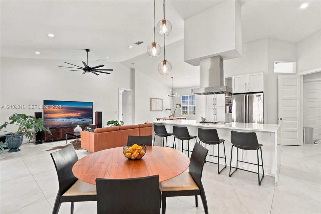 Open-concept interior featuring vaulted ceilings, a large central island with bar seating, custom stainless steel range hood, globe pendant lighting, and light-toned tile flooring