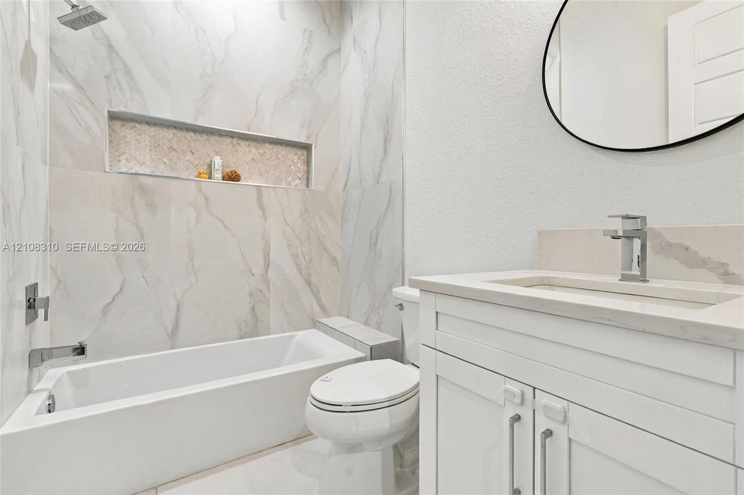 Full-height shower surround with large-format stone-patterned tile, integrated recessed shelving, modern vanity with stone countertop, undermount sink with polished chrome hardware, and oval wall-mounted mirror