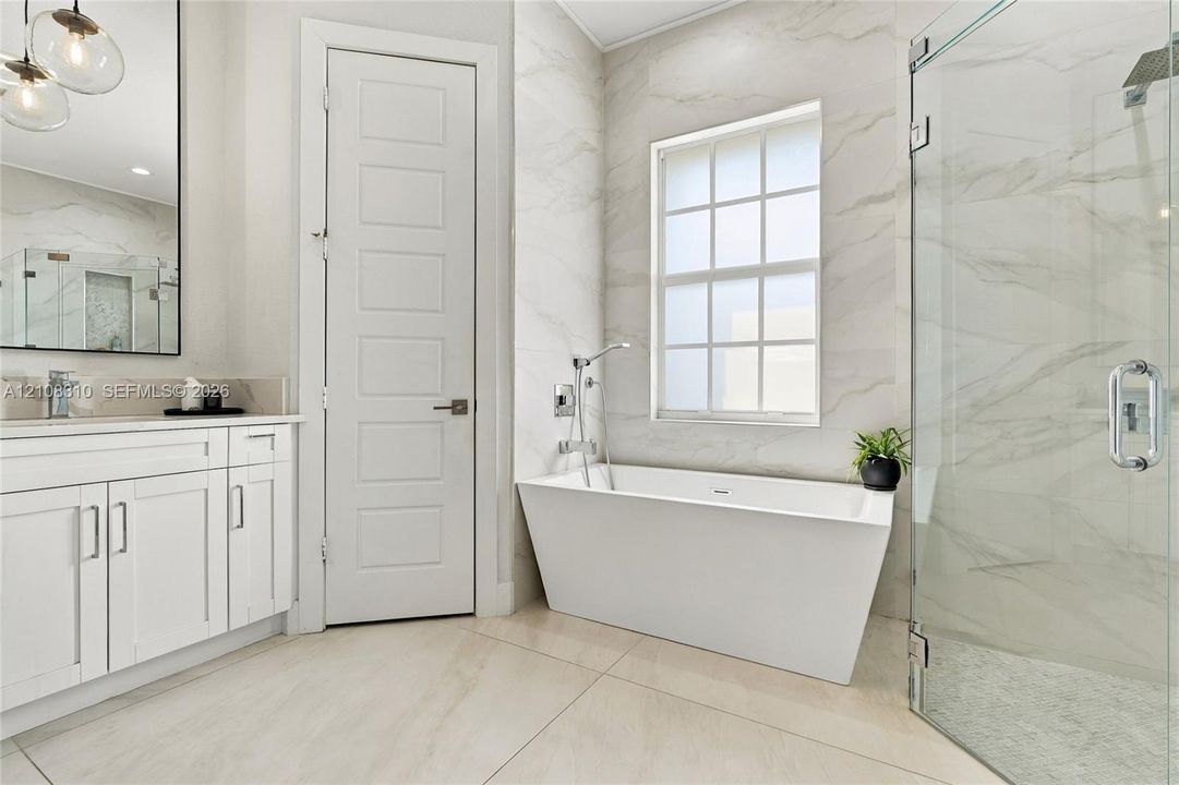 Bathroom featuring a freestanding soaking tub, a frameless glass-enclosed shower, large-format tile flooring, white cabinetry, and natural stone-finish wall tiling