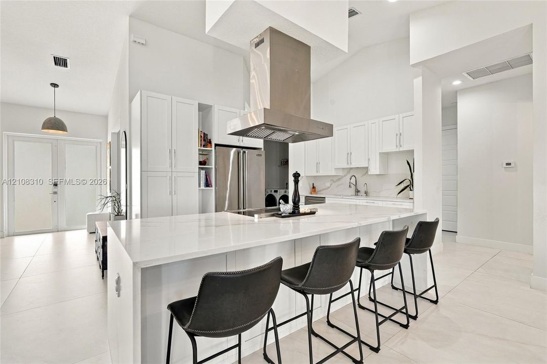 Kitchen featuring white shaker-style cabinetry, a large waterfall-edge island with solid surface countertop, stainless steel appliances, a ceiling-mounted stainless steel range hood, and large-format tile flooring