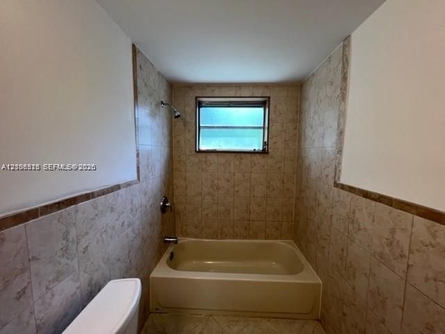 Full-height neutral wall tiling, integrated bathtub, fixed window with exterior exposure, wall-mounted shower fixture, and polished metal hardware