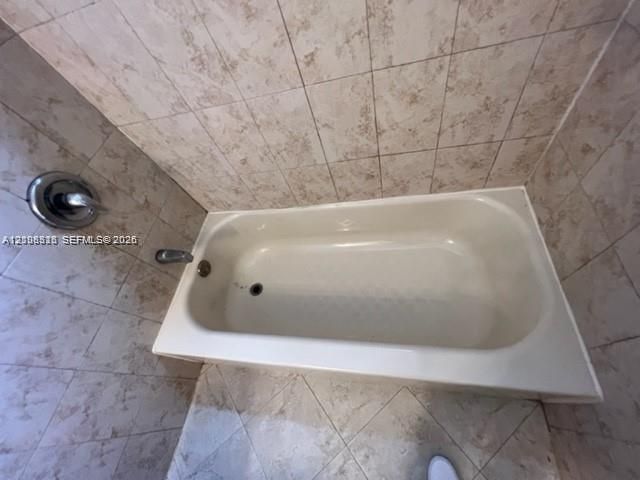 Bathtub enclosed by floor-to-ceiling ceramic wall tile and featuring a chrome-finish shower valve and faucet fixture