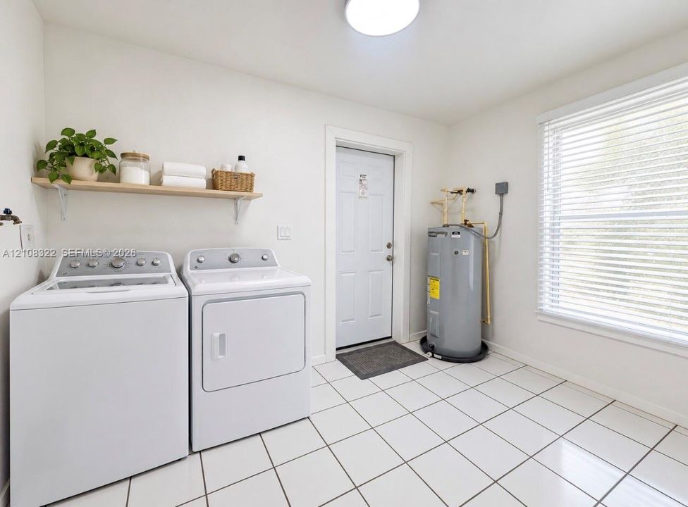 Utility space featuring white ceramic tile flooring, a window with horizontal blinds, a solid-panel exterior door, and a mounted wall shelf