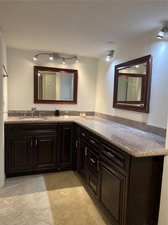 L-shaped vanity featuring dark wood-finish cabinetry, stone-composite countertops, dual framed mirrors, and multi-bulb track lighting