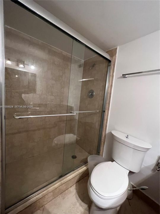 Walk-in shower with a sliding glass door, neutral-toned stone-finish tile surround, and chrome-finish fixtures, complemented by a white ceramic toilet and a wall-mounted metal towel rack