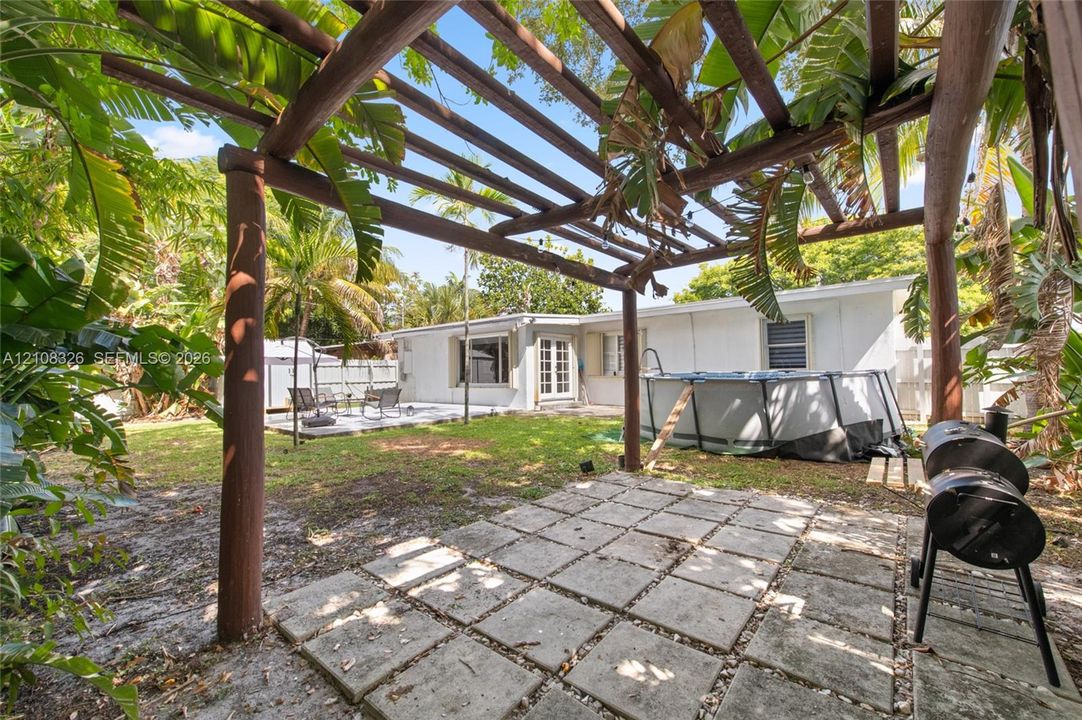Outdoor space featuring a timber pergola, paved patio area, green lawn, and mature tropical foliage