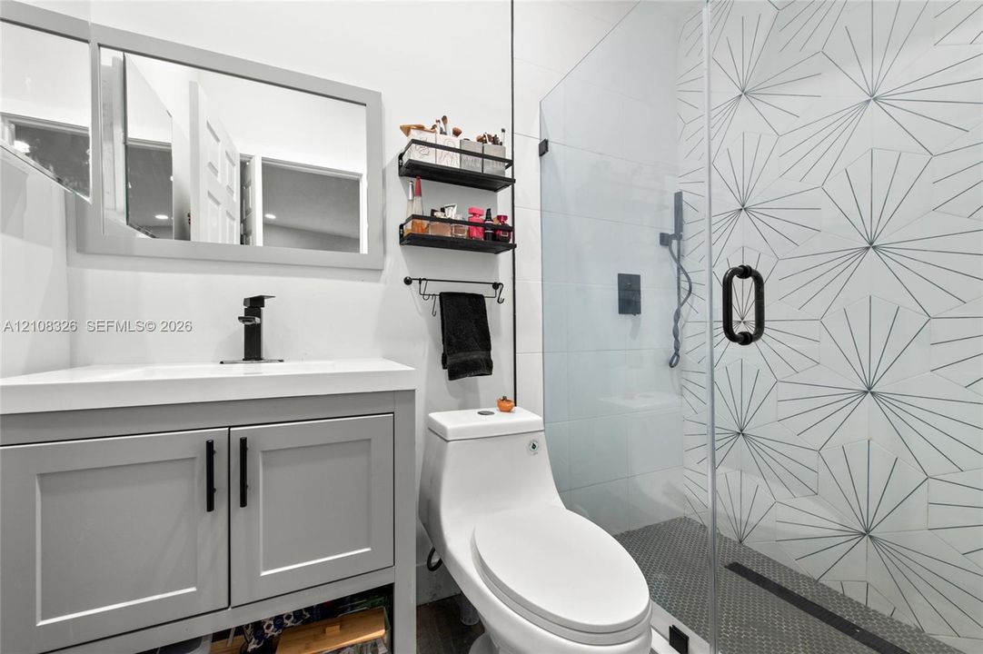 Bathroom featuring a grey vanity with a rectangular sink, matte black faucet, glass-enclosed shower with geometric patterned tile, and a modern one-piece toilet