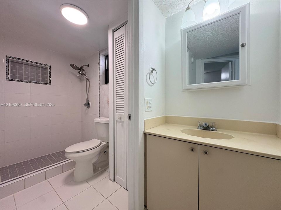Bathroom featuring a walk-in shower with tiled surround and integrated niche, solid-surface vanity countertop with inset sink, medicine cabinet, and louvered-door closet