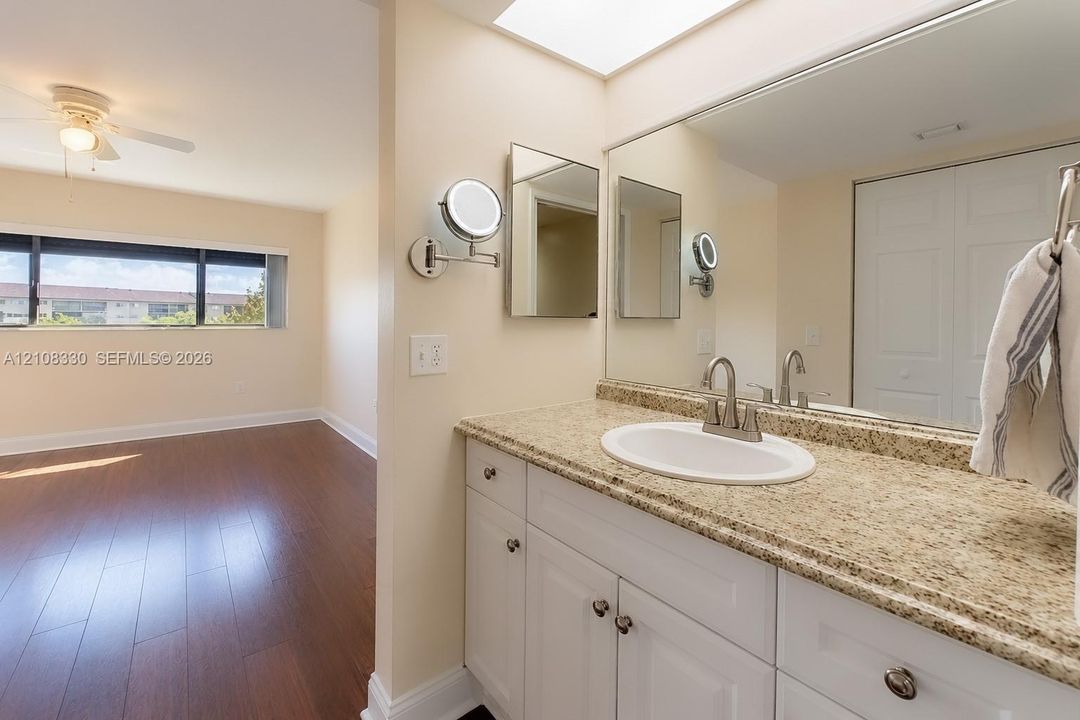 Bathroom vanity featuring white shaker-style cabinetry, neutral stone-finish countertop, undermount sink, large wall-spanning mirror, and adjustable wall-mounted magnifying mirror