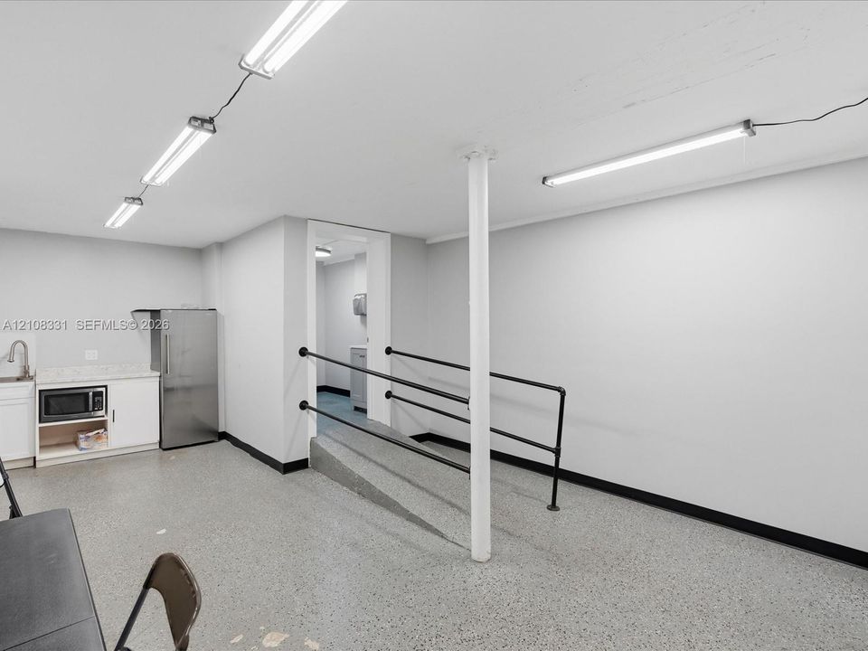 Open interior space featuring speckled flooring, built-in cabinetry with a stone-surface countertop, stainless steel refrigerator, and metal handrails along an inclined ramp